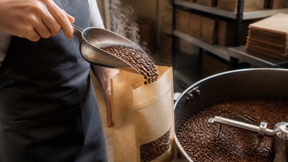 Close-up of freshly roasted medium-brown coffee beans being carefully scooped into a kraft paper bag with a printed roast date label visible, steam still rising slightly, natural warehouse lighting