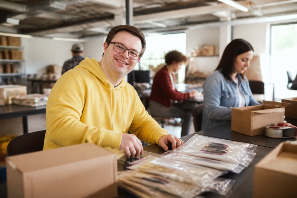 Diverse group of adults with intellectual disabilities working together in a bright, welcoming workplace environment, smiling and engaged in meaningful tasks at their workstations