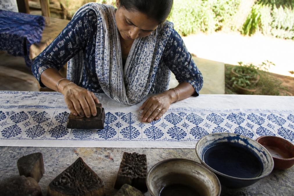 Close-up of a woman's hands block-printing vibrant indigo dye onto a length of organic cotton fabric, with wooden printing blocks and bowls of dye visible on a rustic worktable