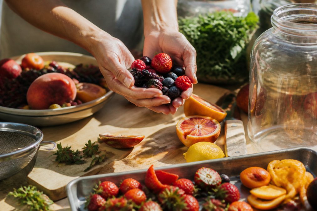 Hands holding fresh colorful farmers market produce including ripe berries, stone fruits, and citrus, being sorted and arranged for fermentation preparation in natural sunlight
