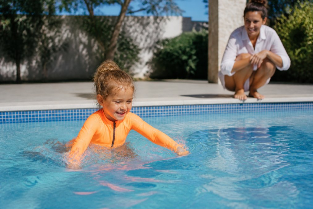 A young child wearing a bright neon orange swimsuit playing in a swimming pool, with a parent watching closely from the poolside, sunny outdoor setting