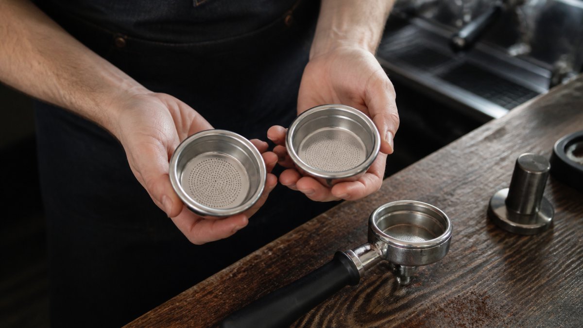 Close-up of a barista's hands holding a polished stainless steel precision espresso basket next to a standard basket on a dark wooden counter, with a portafilter and scattered coffee grounds nearby