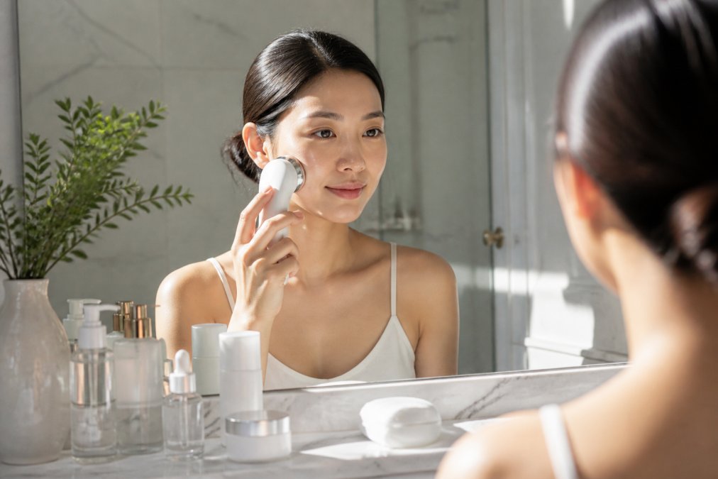 Woman using a handheld facial beauty device on clean skin at home, with serums, toners, and moisturizers arranged on a marble bathroom counter in natural morning light