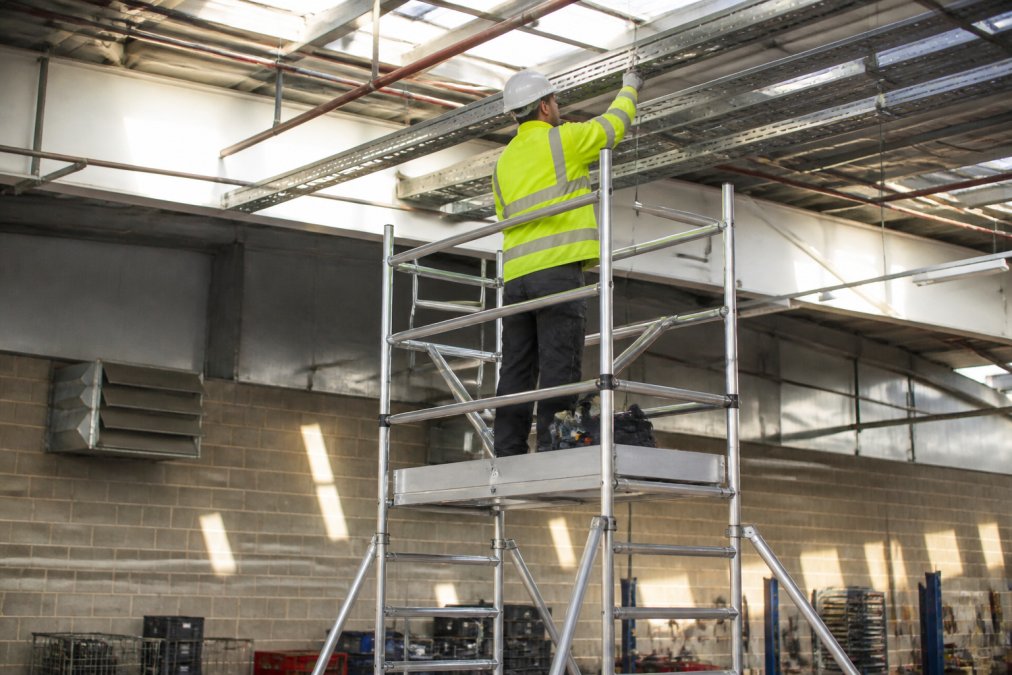 A maintenance technician in high-visibility clothing and a hard hat working from an aluminium mobile tower inside a large industrial warehouse, with tools arranged on the platform and natural light streaming through skylights