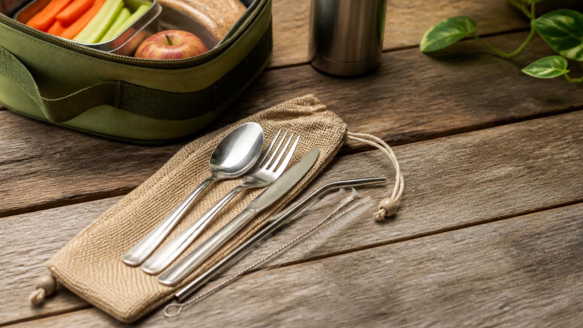 A stainless steel travel cutlery set with a hemp pouch resting on a wooden table beside a packed lunch bag and a reusable water bottle, natural daylight