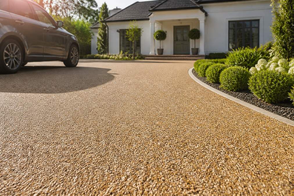 A sunlit residential driveway finished in warm-toned resin bound pebble paving, with a car parked to one side and garden beds bordering the edges, showing the smooth even surface