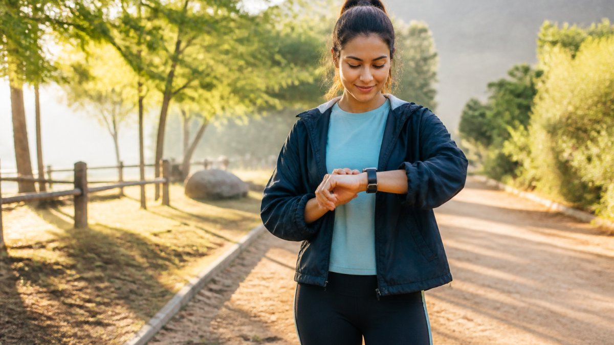 A person in casual workout clothes checking a smartwatch on their wrist while standing outdoors on a walking path, morning light, relaxed expression