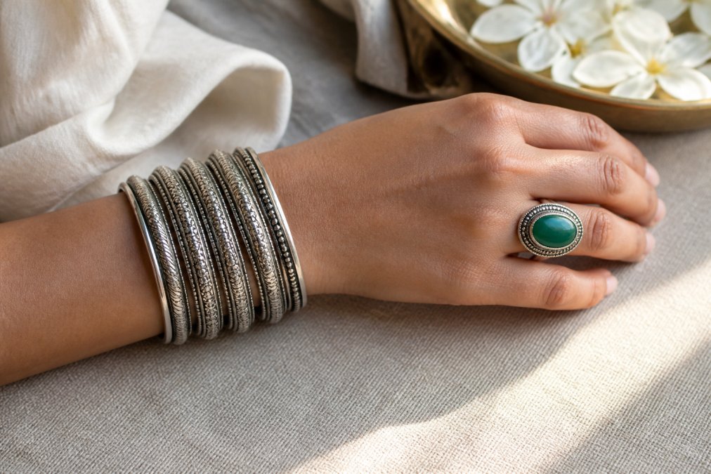 Close-up of a woman's wrist wearing a stack of five silver bangles and a large green aventurine statement ring, resting on a linen tablecloth in soft window light