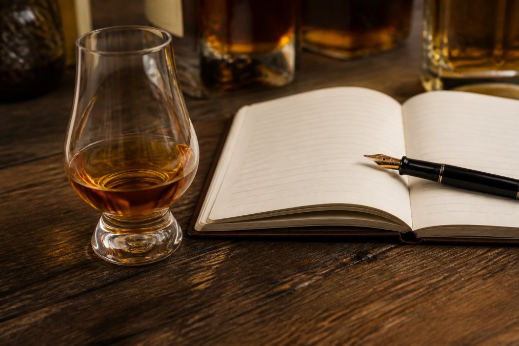 Close-up of a bourbon glass filled with amber whiskey on a dark wooden table, with an open notebook and fountain pen positioned nearby, suggesting active tasting note documentation in warm natural lighting