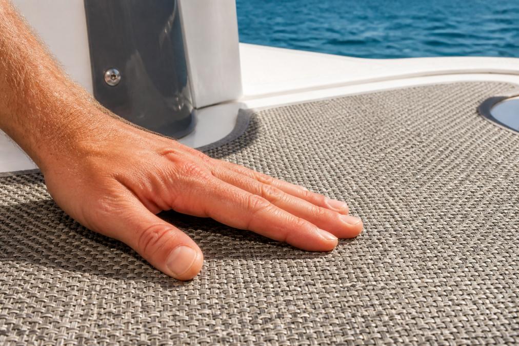 Close-up of a hand pressing on a textured woven vinyl floor on a yacht deck, showing the material's tight weave and non-slip surface under bright sunlight