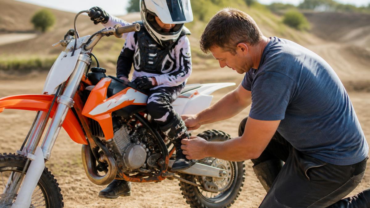 A young rider in full motocross gear sitting on a dirt bike, with an adult parent kneeling to check the fit and buckles of the child's motocross boots on a sunny track