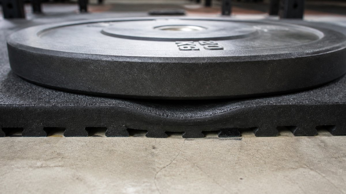 Beskyttelse af undergulv ved tung styrketræning 2 Close-up of interlocking rubber floor tiles with a 20 kg weight plate resting on top, showing compression depth and tile thickness
