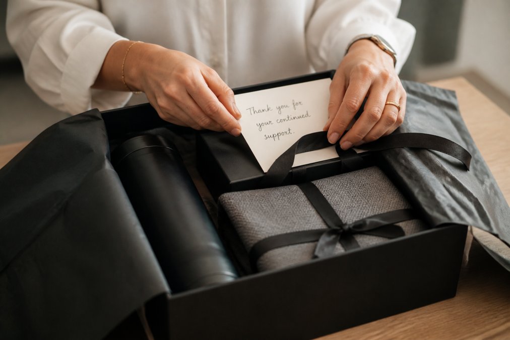 Close-up of hands placing a handwritten card and ribbon into an open premium gift box on a clean desk, with branded tissue paper visible