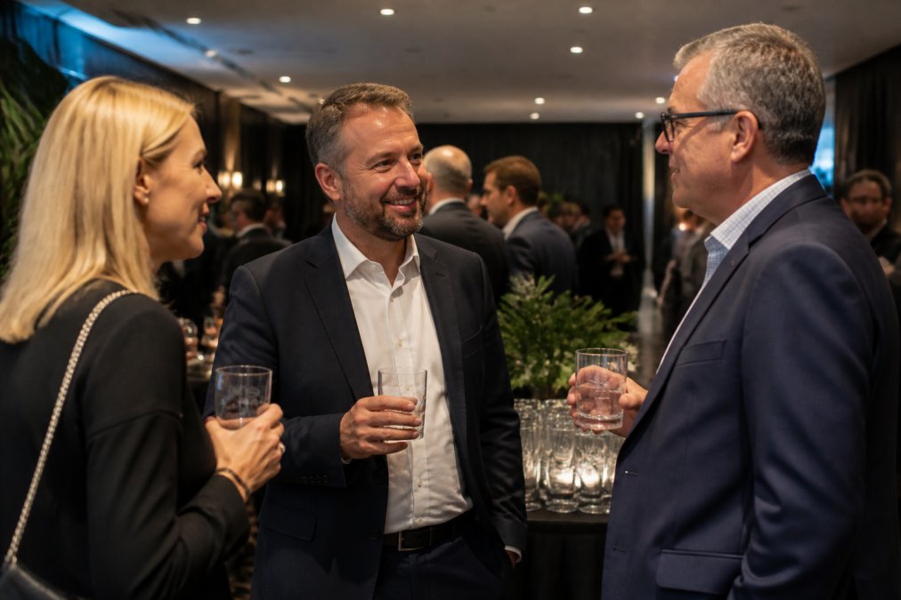 A corporate event or upmarket hospitality setting with branded glassware displayed on a table, guests holding custom-etched glasses in conversation, showing the glassware in real-world use at a professional gathering with elegant venue lighting