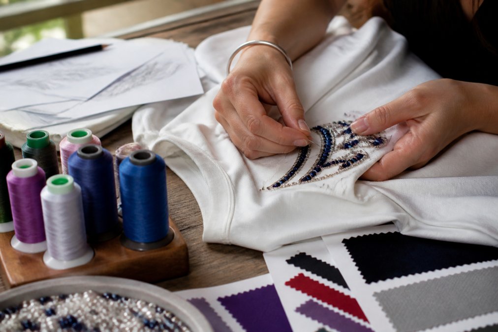 Close-up of hands working on custom embroidery and beading details on a garment, with colorful thread spools, fabric swatches, and design sketches visible on a wooden work table with natural lighting