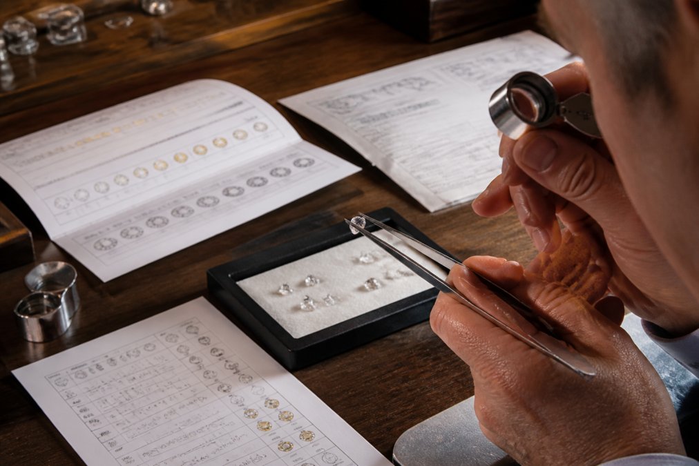 Jeweller examining certified diamonds under magnification with loupe, gemstone certification documents spread across workbench, with colour and clarity reference charts visible under precise studio lighting