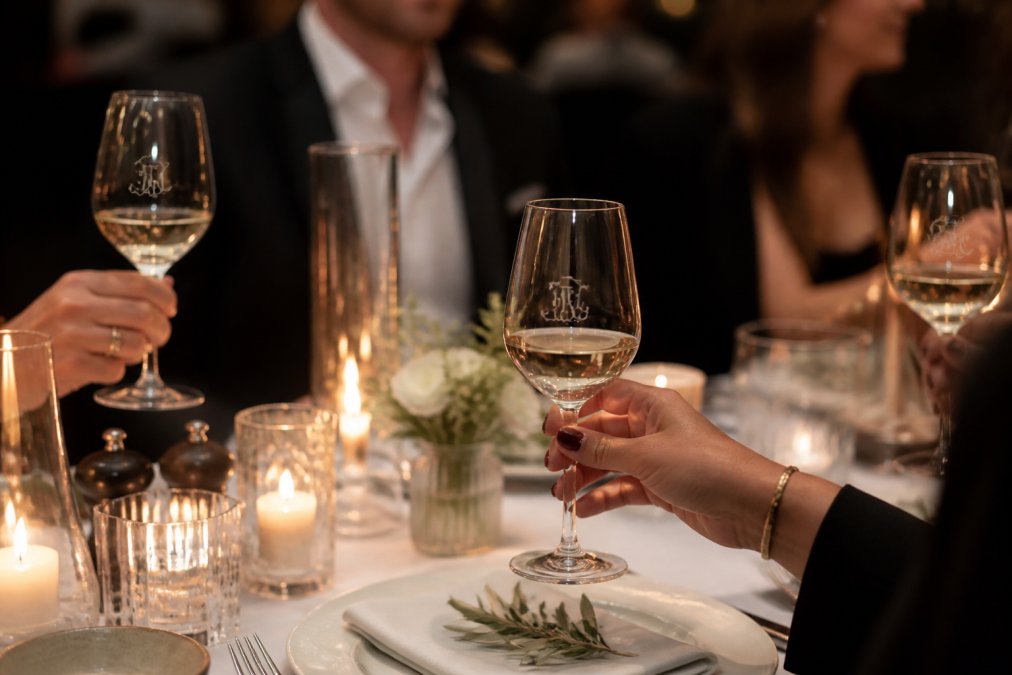 Fine dining table setting with elegant branded wine glasses catching soft candlelight, guests' hands holding glasses with visible etched logo, premium table presentation with glassware as focal point
