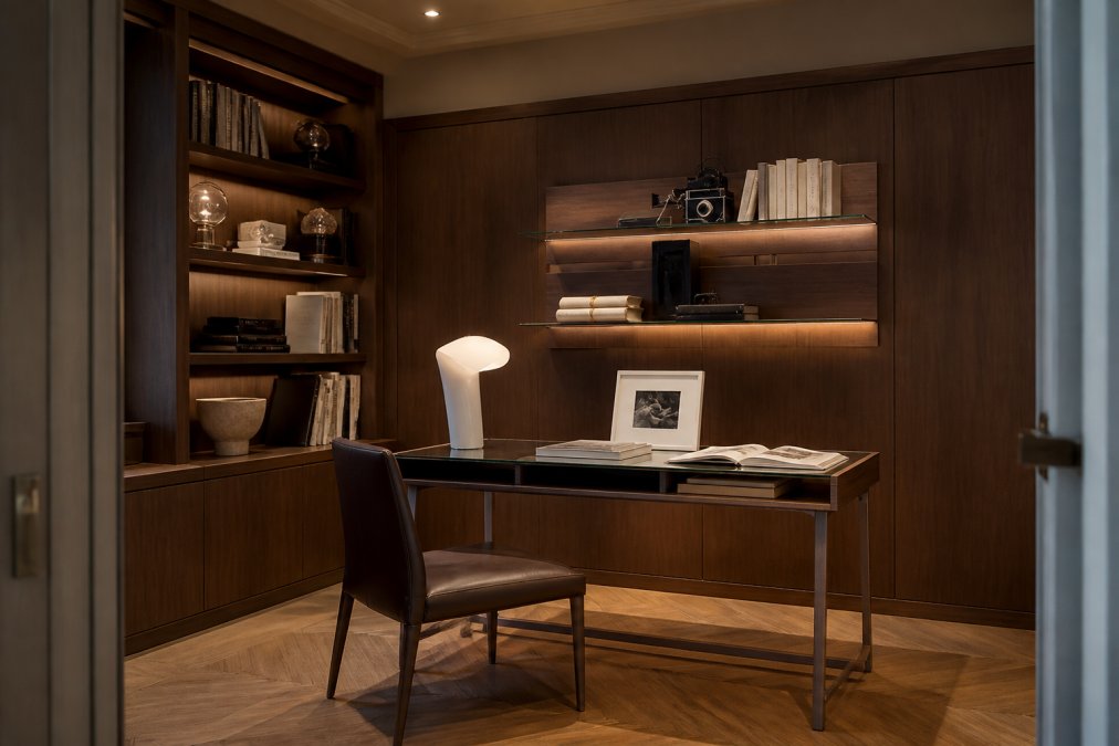 A beautifully appointed home study featuring a solid walnut bespoke writing desk with a tempered glass top, floor-to-ceiling integrated shelving in Canaletto walnut veneer, warm task lighting from a recessed ceiling fixture, and a leather-upholstered chair, photographed in a refined residential interior with parquet flooring