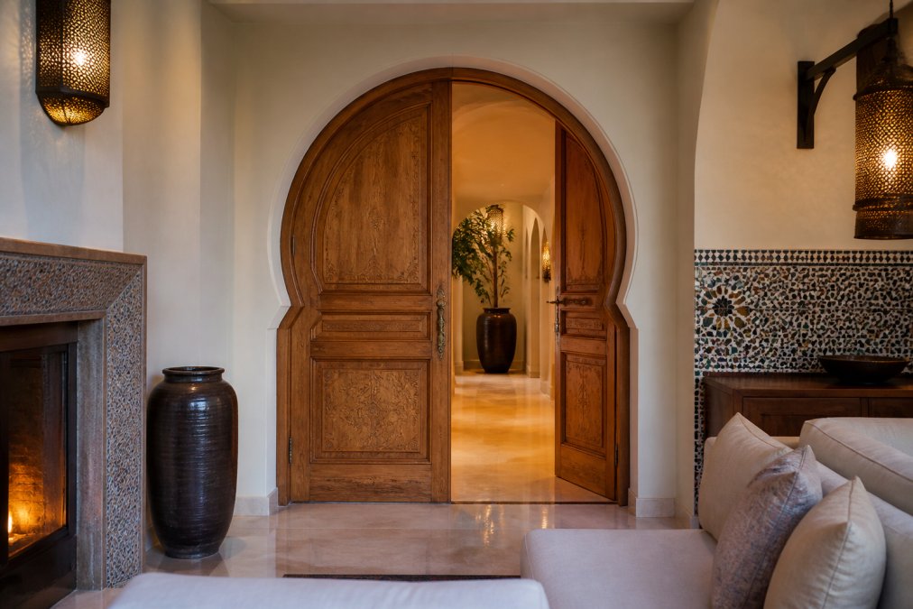 Interior photograph of a luxury modern living space featuring a hand-carved Moroccan wood double door with a horseshoe arch, warm ambient lighting, and subtle zellige tile accents on the wall
