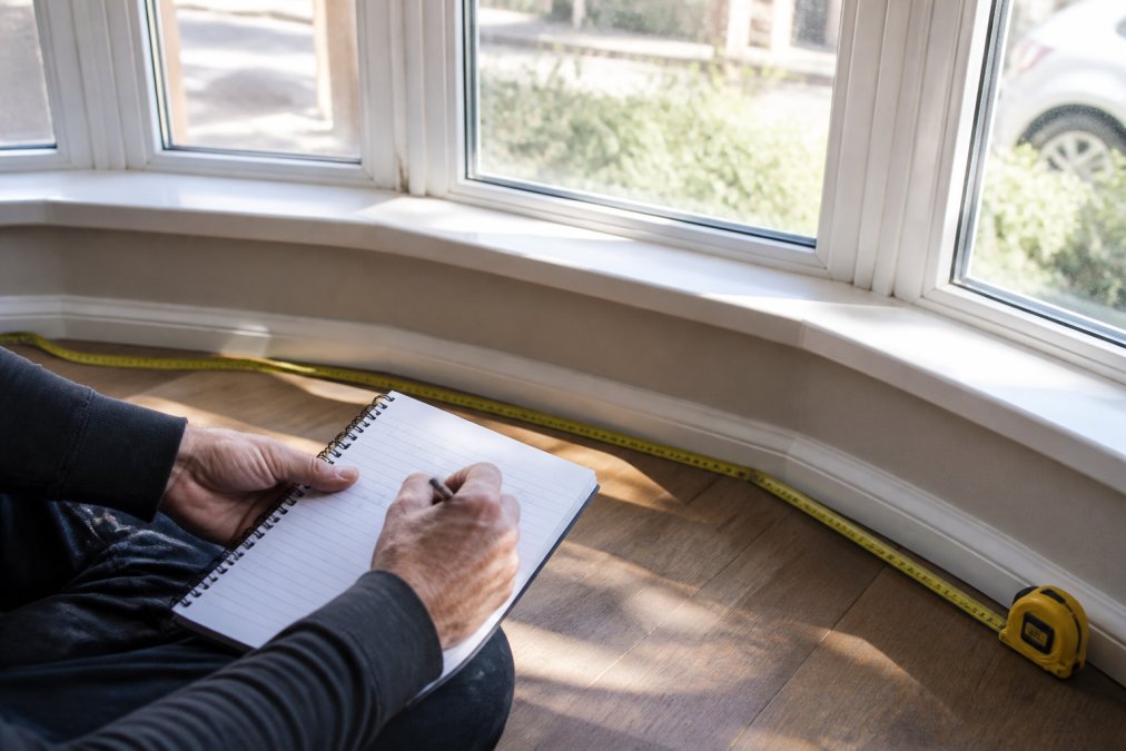 Close-up of measuring tape and notepad recording precise bay window dimensions, natural sunlight streaming through the window panes, professional hand noting measurements on paper with pen