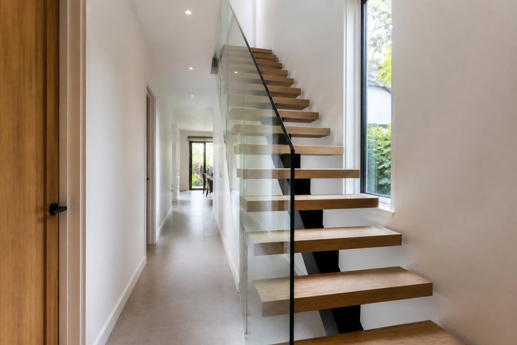 Modern narrow hallway featuring a floating staircase with glass balustrades and open-riser design, natural light streaming through a window, showcasing minimalist aesthetic and visual spaciousness