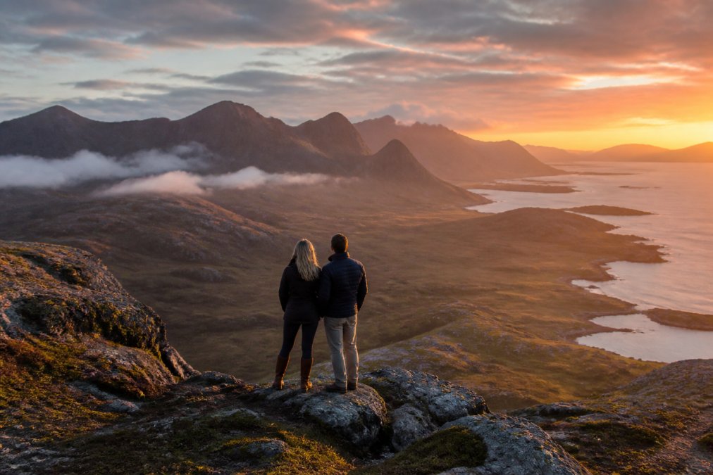 Couple standing on a dramatic clifftop overlooking the Cuillin mountains and rugged Skye coastline at sunset, with golden light illuminating the remote landscape and misty valleys below