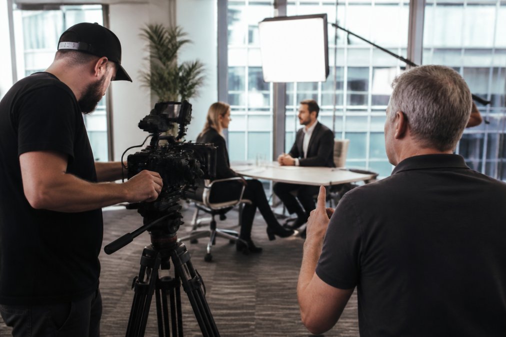 A professional film crew setting up a camera and lighting equipment on a corporate office set, with a director reviewing the shot on a monitor