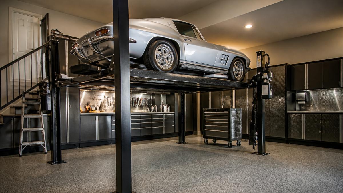 A home garage with a four-post car lift holding a classic sports car at raised height, tools and cabinets along the wall, warm overhead lighting