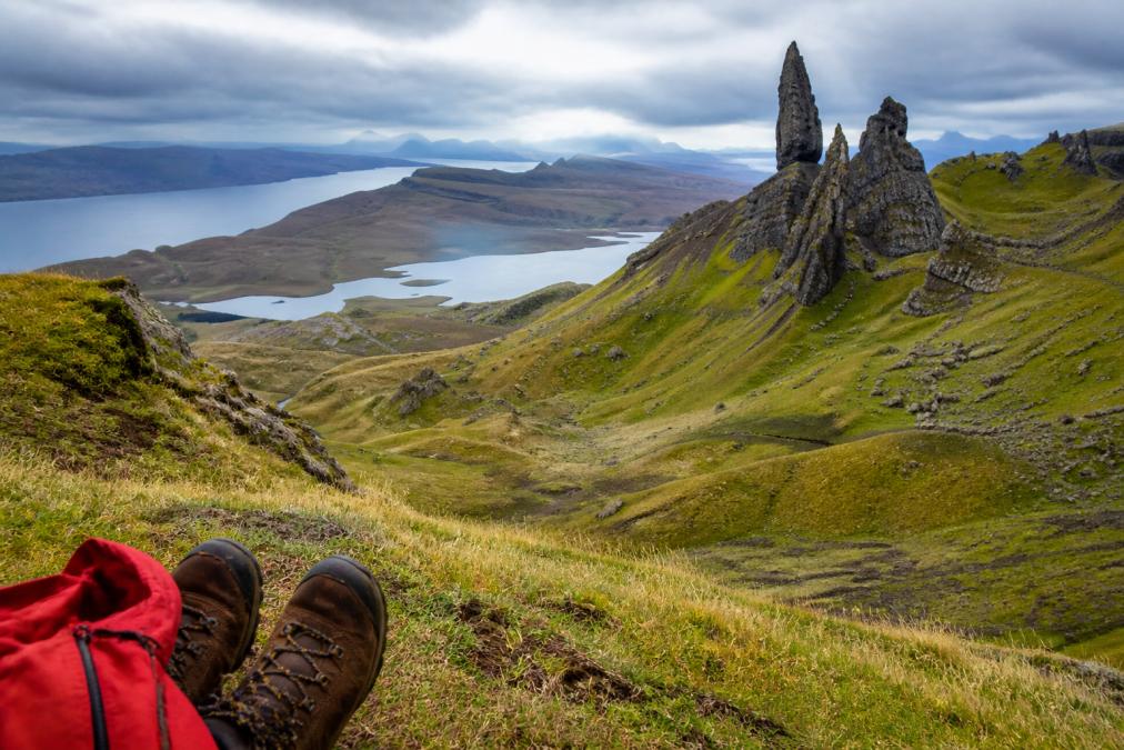 Dramatic landscape of Trotternish Peninsula with steep jagged cliffs rising from moorland, distinctive Old Man of Storr rock pinnacle silhouetted against cloudy sky, hiking boots and waterproof jacket visible in foreground on grassy slope