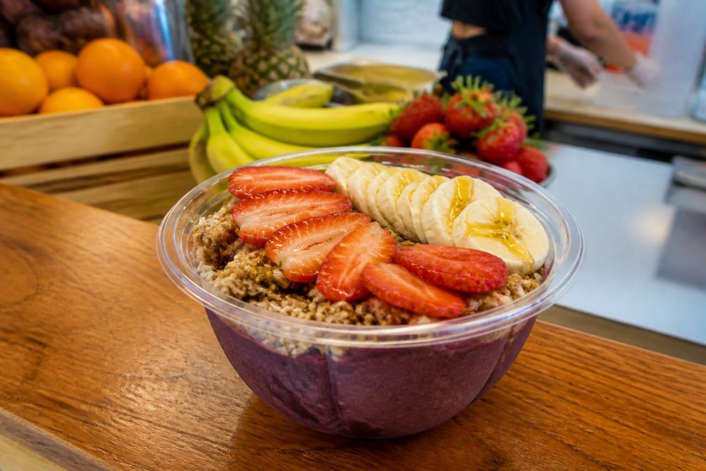 Close-up of a freshly made acai bowl topped with granola, sliced strawberries, banana, and a honey drizzle on a wooden counter, with a blurred juice bar background showing fresh fruit