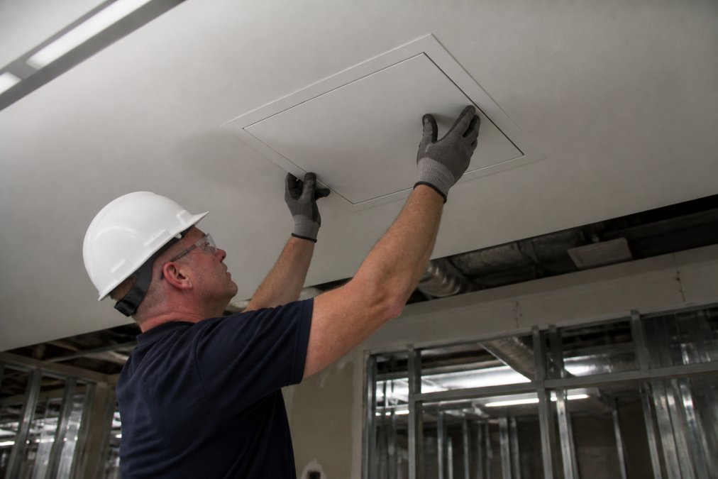 A contractor in a hard hat and work gloves installing a flush steel access panel into a drywall ceiling in a commercial building interior, fluorescent overhead lighting, exposed framing visible in background