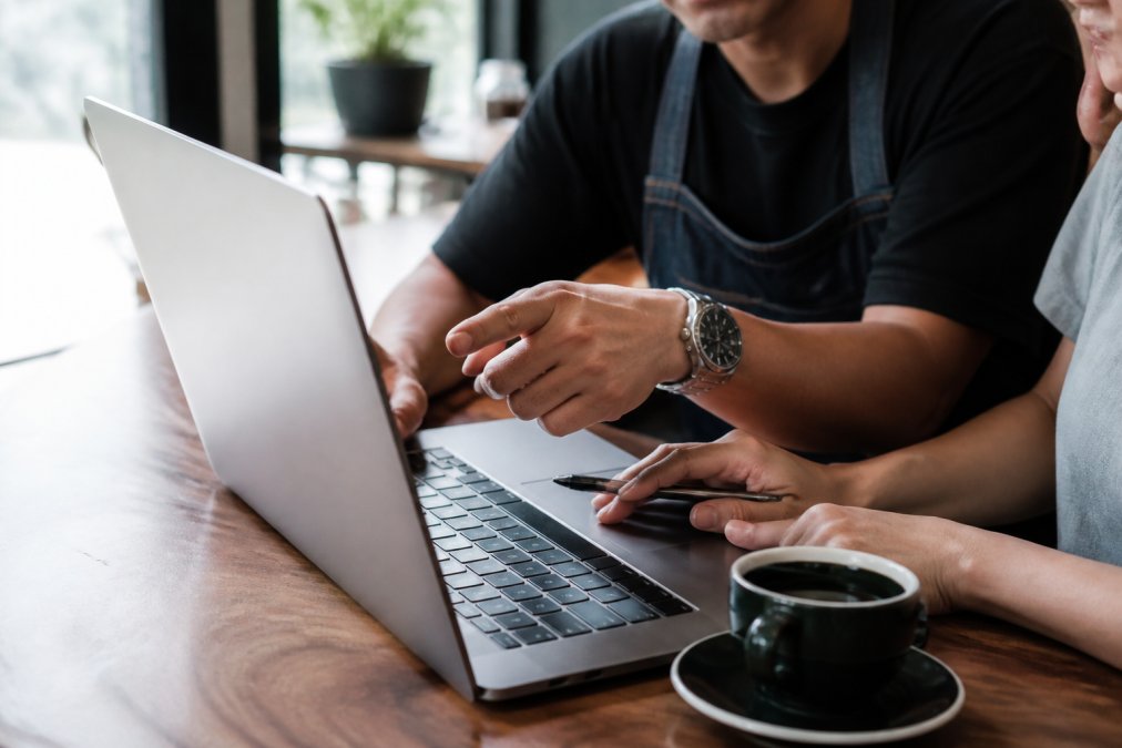 Close-up of hands pointing at a laptop screen displaying financial metrics and restaurant KPI dashboard with graphs, coffee cup beside the keyboard in a professional workspace with natural light