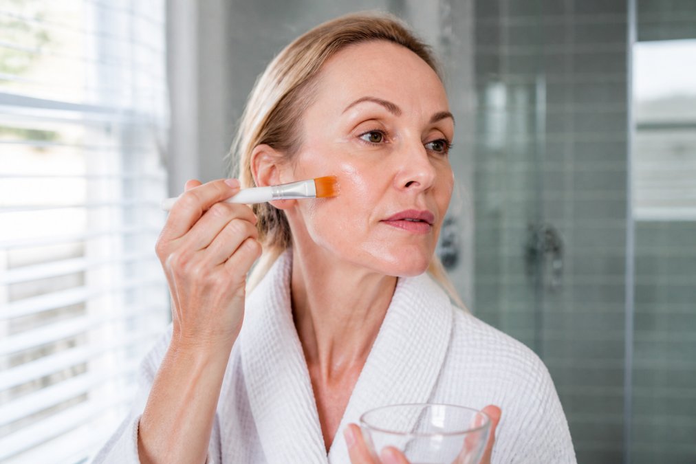 Close-up of mature woman's hands applying chemical peel solution to clean facial skin with a brush, showing proper technique in a bright bathroom with natural lighting