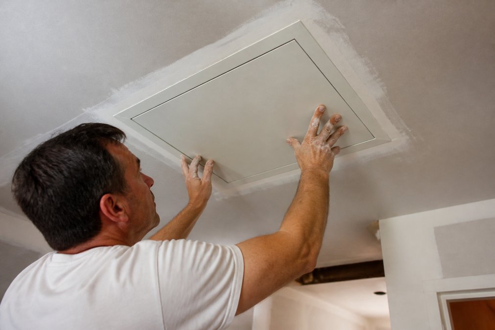 Professional contractor installing a flush-mount access door in a drywall ceiling, showing the paintable frame and proper flange alignment with drywall tape and joint compound