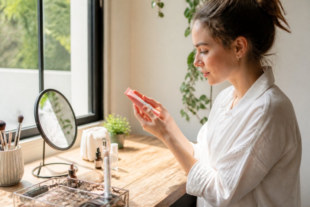 Woman reading ingredient labels on lip product packaging in natural daylight, examining the back of a cosmetic tube closely while holding it up to the light