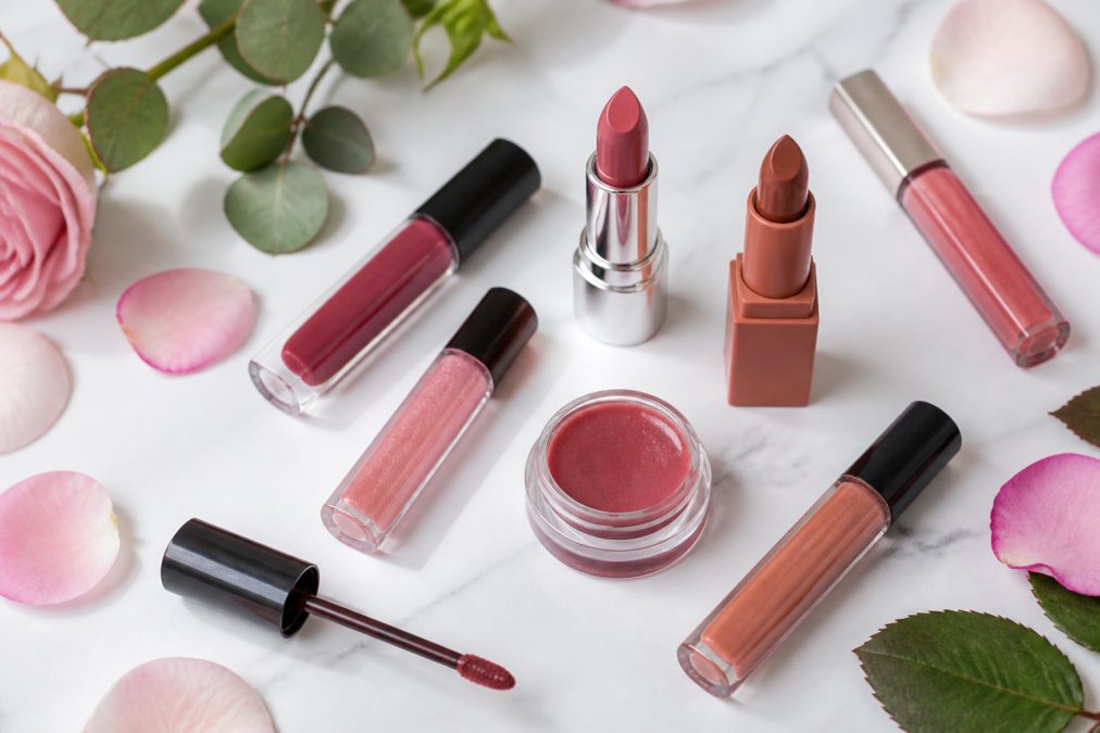 Flatlay of diverse lip beauty products including glosses, lipsticks, and stains arranged on a clean white surface with fresh botanical elements like rose petals and green leaves