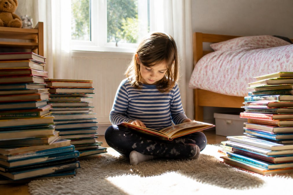 Child sitting cross-legged on a bedroom floor surrounded by colourful stacks of pre-loved children's books, reading intently with natural sunlight streaming through a window