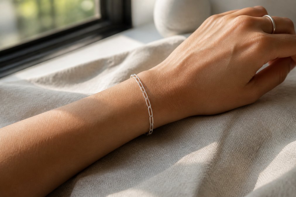 Close-up of a woman's wrist and hand wearing a delicate sterling silver chain link bracelet, soft natural window light highlighting the metal's shine against a neutral linen background