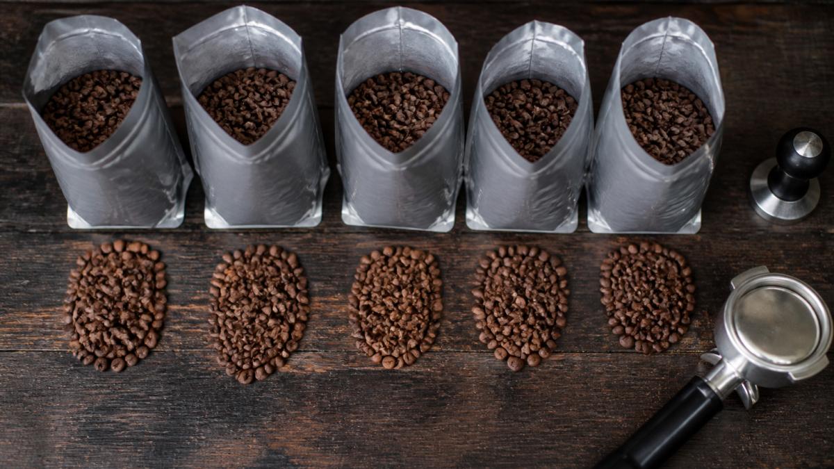 Five open bags of roasted coffee beans arranged on a dark timber bench with a portafilter and tamper nearby.