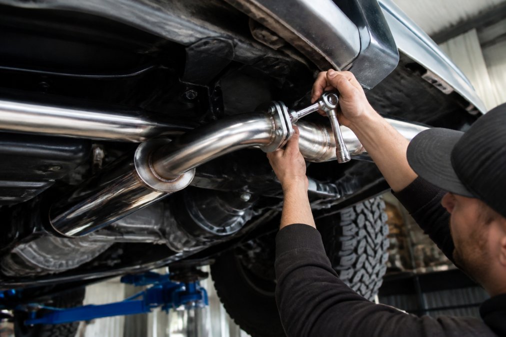 Close-up of a stainless steel performance exhaust system being fitted to a 4x4 ute in a well-lit workshop, with a mechanic's hands tightening flanged joints using a ratchet