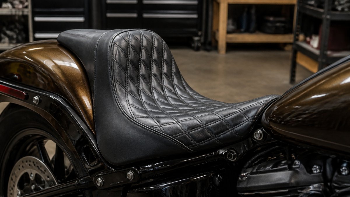 A close-up photograph of a handcrafted leather motorcycle seat mounted on a Harley-Davidson, showing diamond stitching and a contoured fastback shape, with a workshop or garage setting in the soft-focus background