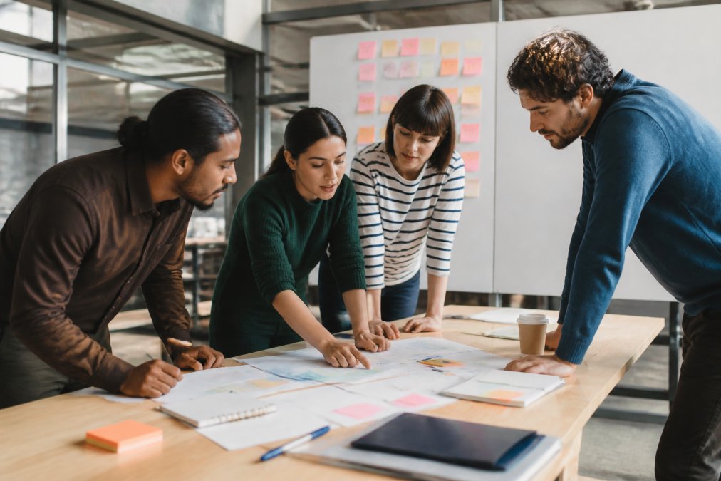Cross-functional software development team gathered around a desk reviewing sprint progress on a large monitor in a bright, modern office environment