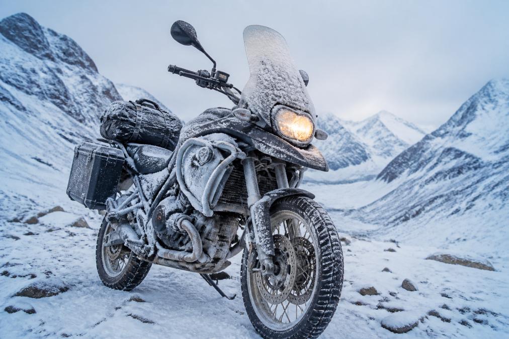 Touring motorcycle parked in snow-covered mountain terrain with frost coating engine components and headlight, illustrating extreme cold-weather touring conditions