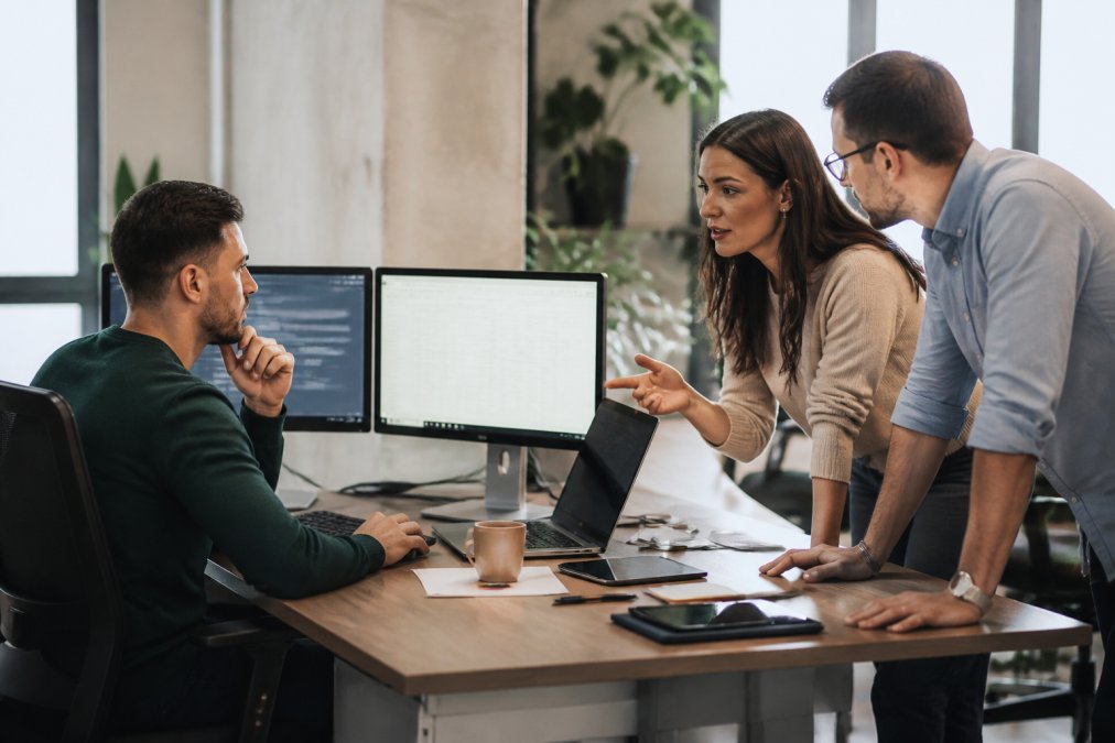Business owner reviewing workflow automation on a computer screen with team members discussing strategy around a modern office desk with multiple monitors in a bright, modern office setting
