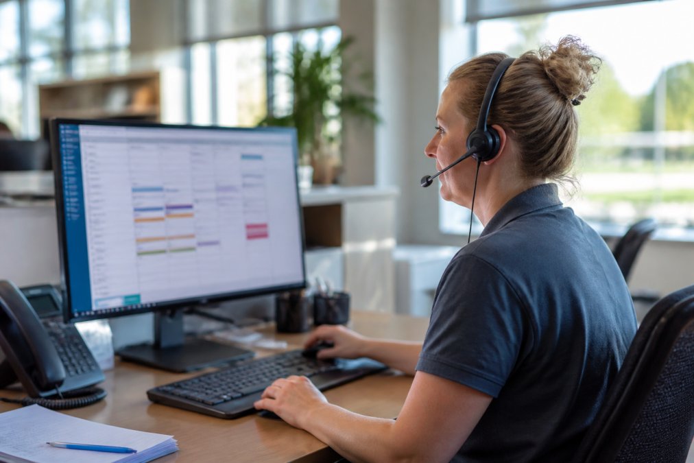 Dispatcher wearing a headset at a desk reviewing a job scheduling calendar on a large computer monitor in a bright home services office with natural light through windows