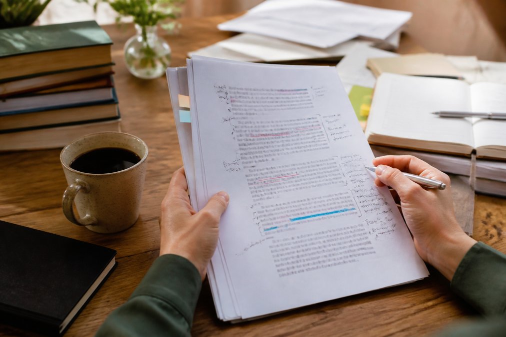 Close-up of hands holding printed manuscript pages with character notes and annotations scattered on desk, coffee and pen nearby