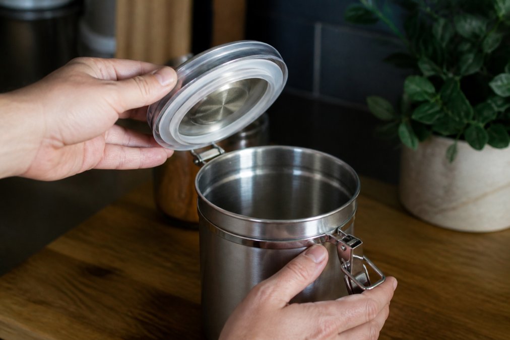 Hands carefully opening a stainless steel coffee canister with a visible silicone gasket seal, showing the lid mechanism and interior vacuum seal detail in natural kitchen lighting