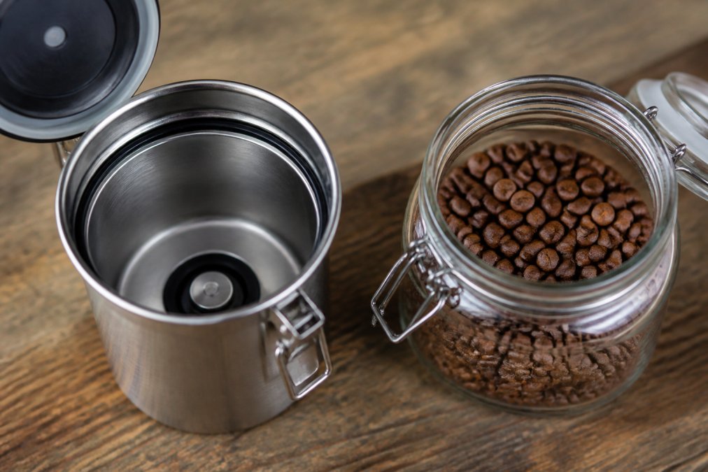 Close-up of an open stainless steel coffee canister showing the vacuum seal mechanism next to an open glass container filled with whole coffee beans, both displaying their interior clarity and seal integrity in natural light