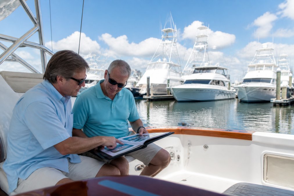 A yacht broker and a buyer reviewing a vessel comparison document on a tablet aboard a sportfish yacht at a marina, with other yachts visible in the background