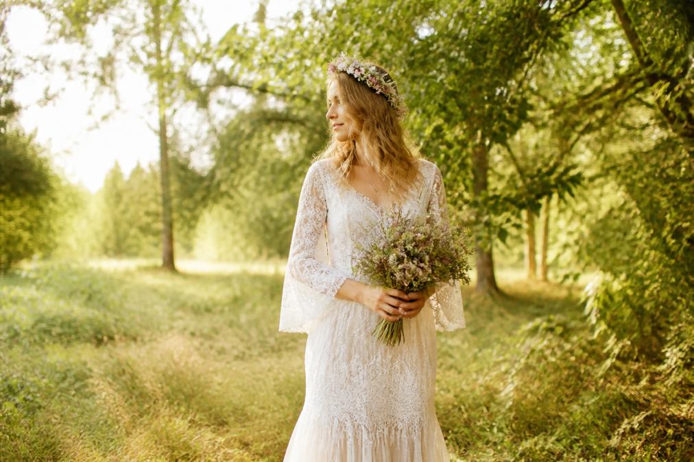 A bride wearing a bohemian lace wedding dress with a flower crown, standing in a sunlit outdoor setting with natural greenery, holding a simple bouquet of wildflowers